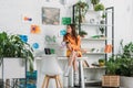 Girl sitting on desk and holding paper cup in room decorated with green plants and paintings on wall