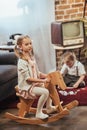 girl sitting on rocking horse and looking at camera and little brother playing with domino tiles behind at home Royalty Free Stock Photo