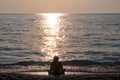 Girl sitting on sandy beach next to sea and enjoys watching sunset, reflection of sun rays over sea. Silhouette of woman by ocean. Royalty Free Stock Photo