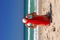 Girl sitting on sandy beach in the sun adjusting hat. Blue sky, blue sea red scarf. Spain. Royalty Free Stock Photo