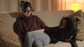 A girl is reading a tablet and sitting on a sofa while caressing a dog.