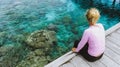 Girl sitting on a wooden jetty near Mansuar island in Raja Ampat. Beautiful colorful corals visible in transparent