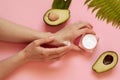 Girl smears cream hands. Two halves of avocado with leaves, a jar of cream and a leaf of fern on a pink background. Natural Organi