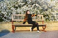 Girl on smiling face sits on bench, sakura on background, defocused. Girl relaxing while walk in park near cherry blossom. Fashion