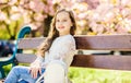 Girl on smiling face sits on bench, sakura tree on background, defocused. Cute child with long beautiful hair enjoy