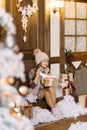 A girl in a smoky pink hat, fur vest, dress and eels sits on the porch of a house decorated for Christmas and holds a gift