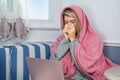 A girl sneezes wrapped in a blanket and uses a laptop