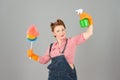 Girl with soft colorful duster and green spray bottle on grey background. Denim girl in orange gloves with brunette curls