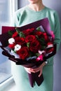 A girl standing near the window in a sea-colored dress holds in her hands a burgundy bouquet with red roses with white flowers and Royalty Free Stock Photo
