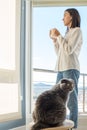 Girl standing by window drinking coffee with her pet gray cat. The tranquility of home with the whole family comfortably