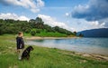 Girl stands near dog of Rottweiler breed in meadow next to lake, against hilly valley with spruce forests