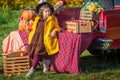 A girl stands near a red retro car loaded with boxes of vegetables with an apple in her hand against the backdrop of rural autumn