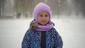 A girl stands in a park in winter and brushes off snowflakes. A girl in a jacket and hat stands under heavy snowfall and