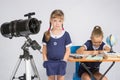 A girl stands at a telescope with his hands in his pockets, another girl having fun sitting at the table and writes