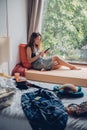 Woman tourist sitting near window using mobile phone in room with mess in hotel