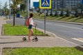 A girl in the summer in the city crosses the road with a scooter, casual wear denim shorts, a white shirt and a pink Royalty Free Stock Photo