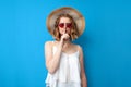 Girl in a sun hat and glasses shows a gesture of silence on a blue isolated background, woman hides a secret in summer clothes, Royalty Free Stock Photo