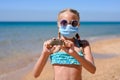 A girl in sunglasses and a medical mask stands on the beach and shows with two hands a stone with the inscription Covid-19.
