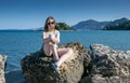 A girl in sunglasses is sitting on rocks on the beach with a small island in the background on a sunny summer day Royalty Free Stock Photo