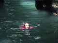 A girl in a sunlit face, closing her eyes, floats on emerald water between the rocks