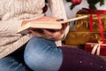 Girl in sweater holding book in hands and Christmas tree and gift box in background Royalty Free Stock Photo