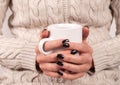 Girl in sweater holding cup of tea in her hands with black nails, close up