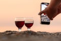 A girl takes a picture of a glass of wine on her phone at sunset on the sea