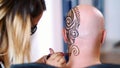 A girl, tattoo master, mehendi artist makes drawing of henna tattoo on scalp of bald Caucasian man, shoulder, neck.The Royalty Free Stock Photo