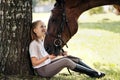 Girl teenager jockey sits in a green clearing under a tree. Feeds a horse an apple and strokes it. Royalty Free Stock Photo