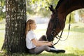 Girl teenager jockey sits in a green clearing under a tree. Feeds a horse an apple and strokes it. Royalty Free Stock Photo