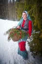 Girl in thick coat and a red sash with basket of fir branches and berries in cold winter day in forest. Medieval peasant