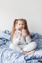 A girl with two ponytails in soft gray pajamas sits on a bed and bites a bar of chocolate in foil