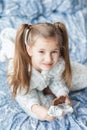 A girl with two ponytails in soft gray pajamas sits on a bed and bites a bar of chocolate in foil