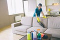 Girl in unform stands behind sofa and cleans it with small vacuum cleaner. There are bottles with different liquid and