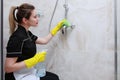 Girl in uniform cleaning the faucet in the shower stall. Napkin and spray in hand.The bathroom is decorated with marble