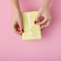 Girl unties a ribbon bow on a gift box with hands, women with present wrapped in decorative paper on pink background, top view,