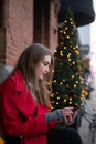 Girl uses her smartphone at Christmas. Young woman sitting on a bench near a Christmas tree. Blurred Christmas lights with bokeh Royalty Free Stock Photo