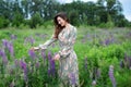 Girl walking on field on summer with wildflowers. Woman in long dress stands in blooming purple lupine field. Beautiful girl with