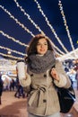 Girl Walking with a hot drink on christmas decoration lights street. Young happy smilin Woman wearing stylish knitted scarf, coat