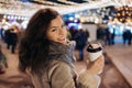 Girl Walking with a hot drink on christmas decoration lights street. Young happy smilin Woman wearing stylish knitted scarf, coat