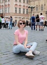 The girl in a pink undershirt and in dark glasses sits on stone blocks at Old Town Square in Prague.