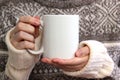 Girl in a warm sweater is holding white mug in hands.