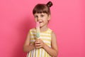 Girl with water ice cream looking directly at camera, wearing white and yellow summer dress, posing against pink wall, licking Royalty Free Stock Photo