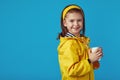 Girl wears raincoat, holds takeaway cup with tea and smiling against blue