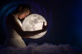 Girl in white air dress with the moon. Beautiful tender girl on the night sky with bright stars and soft clouds hugs the moon.