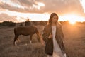 A girl in a white airy dress stands next to a horse and poses in the forest at sunset, like in a fairy tale. Well Royalty Free Stock Photo