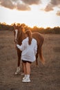 A girl in a white airy dress stands next to a horse and poses in the forest at sunset, like in a fairy tale. Well Royalty Free Stock Photo