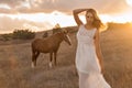 A girl in a white airy dress stands next to a horse and poses in the forest at sunset, like in a fairy tale. Well Royalty Free Stock Photo