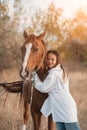 A girl in a white airy dress stands next to a horse and poses in the forest at sunset, like in a fairy tale. Well Royalty Free Stock Photo