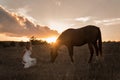 A girl in a white airy dress stands next to a horse and poses in the forest at sunset, like in a fairy tale. Well Royalty Free Stock Photo